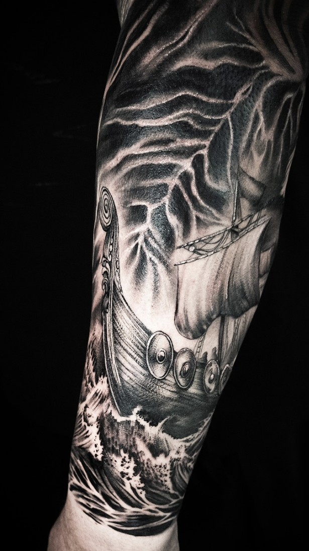 Black and white tattoo of a ship and waves on a person's arm against a black background