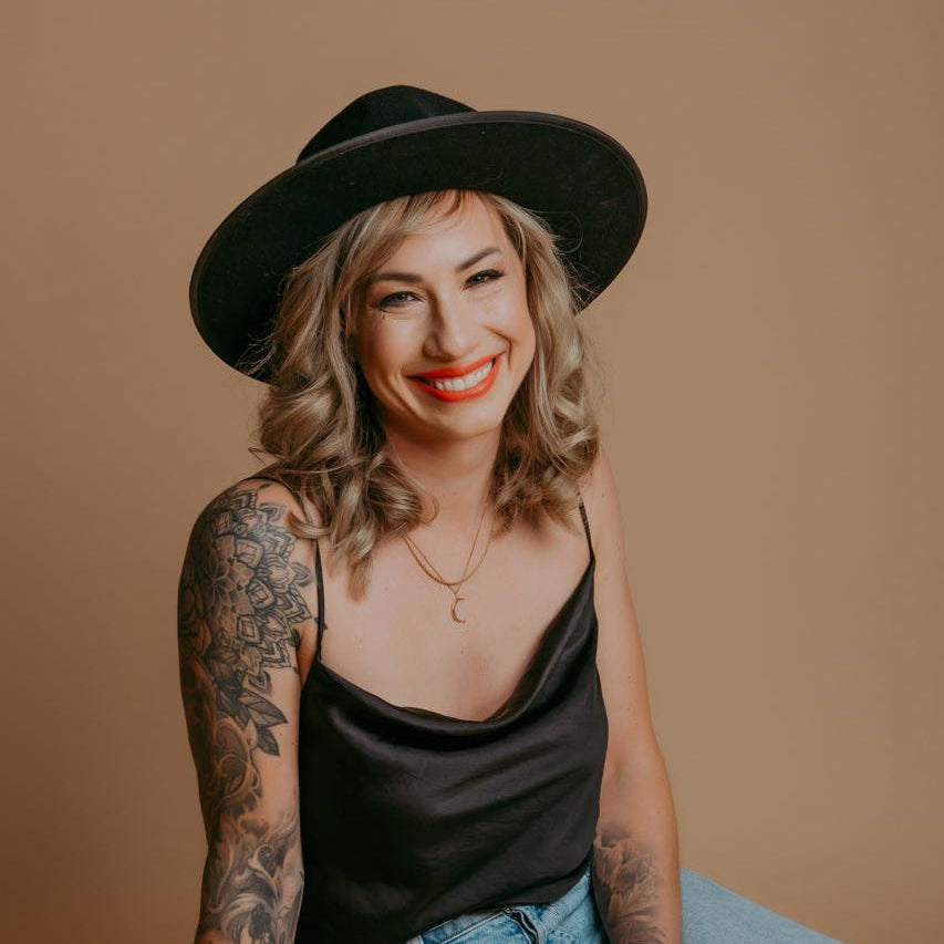 Yvonne a Professional Tattoo Artist at Sundog Studios in Airdrie Alberta wearing a black hat and black top sitting with tattoo sleeves