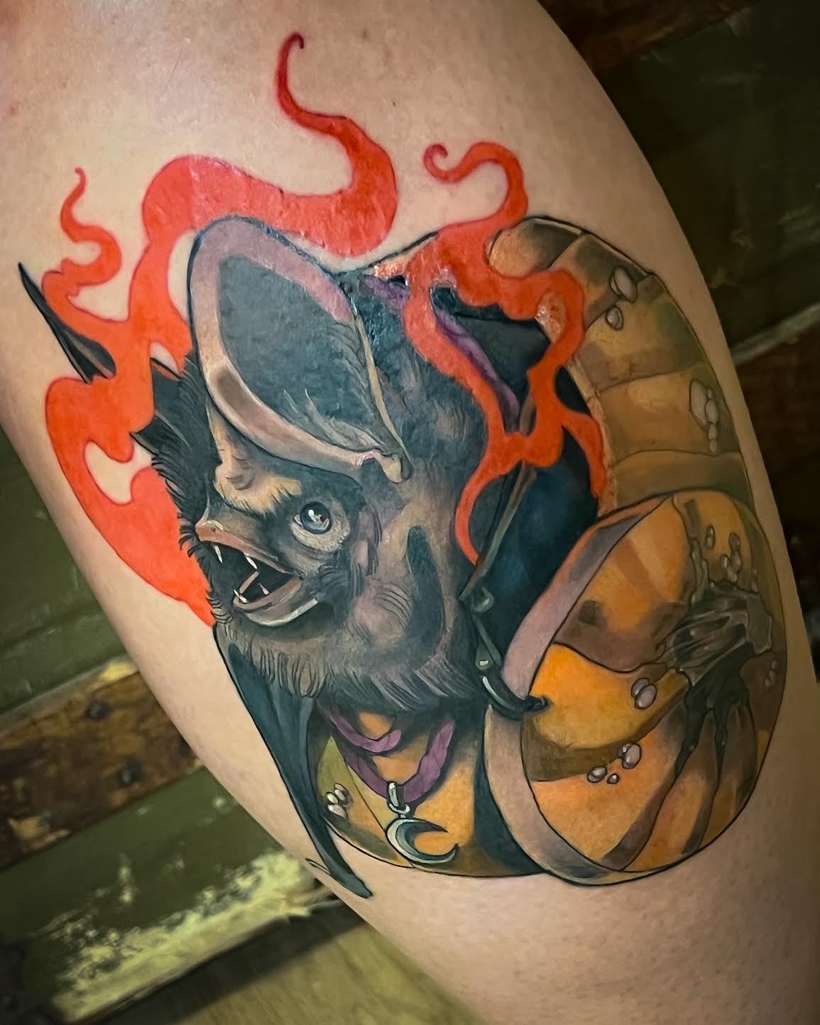 A close-up of a tattoo on human skin showing a stylized bat with bright red flames and a shield, photographed in a real environment.