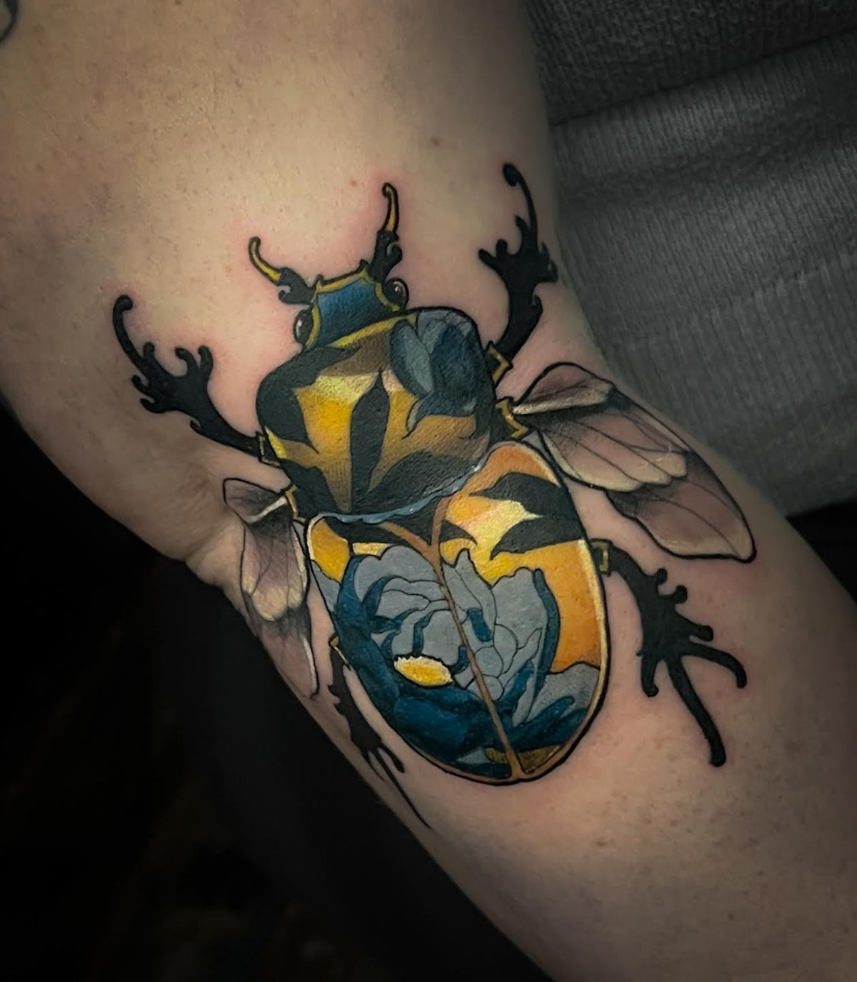 A close-up of a colorful tattoo on a person's upper arm, depicting a beetle with blue, yellow, and black floral details and black legs and wings.
