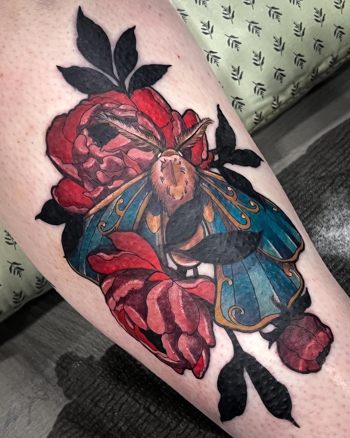 A close-up of a freshly tattooed forearm featuring a large red rose, a blue and gold butterfly and black leaves on light skin.