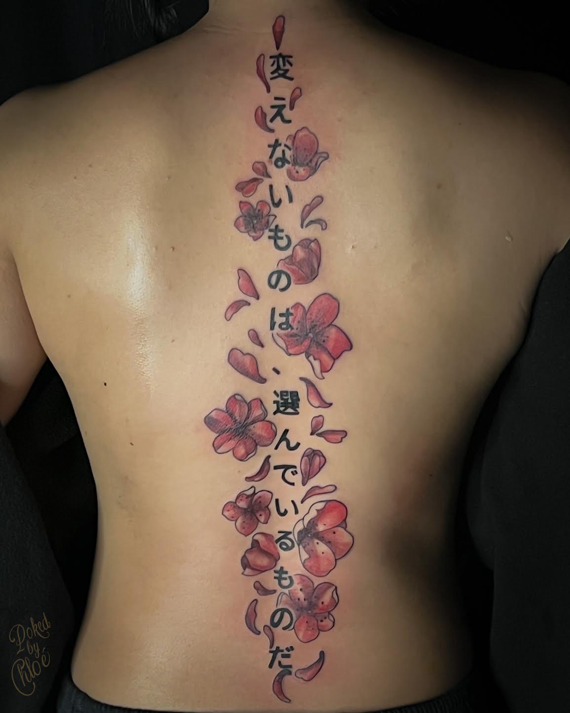 A back tattoo featuring vertical black Japanese calligraphy surrounded by red and pink cherry blossom flowers and petals, shown on a person's bare back.