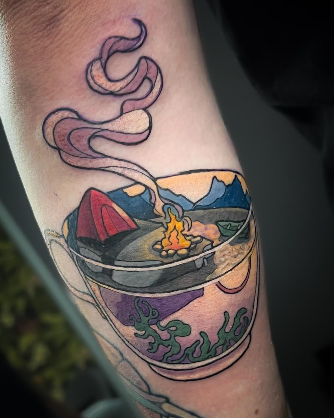 A close-up of a tattoo on a person's forearm featuring a teacup filled with a camping scene, including a red tent, mountains, a campfire, and pink steam.