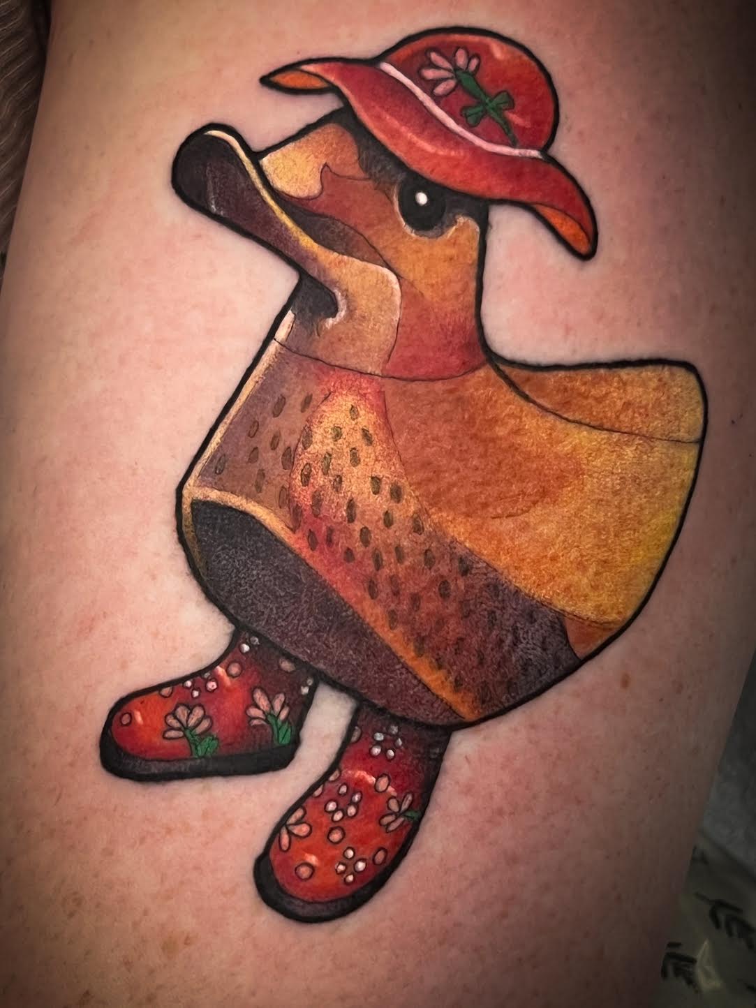 A colorful tattoo of a duck wearing a red hat and red floral boots, shown on human skin.