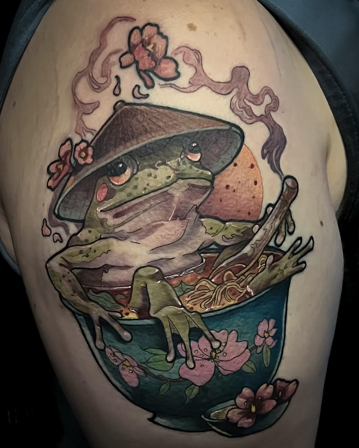 A large, colorful tattoo on a person's upper shoulder featuring a green frog wearing a conical hat and sitting in a bowl of ramen with pink cherry blossoms.