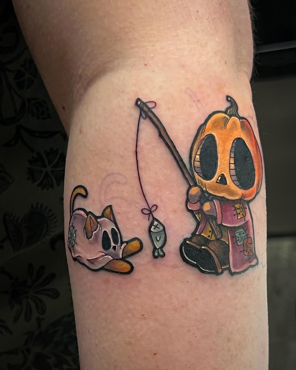 A close-up of a forearm featuring a colourful tattoo of a skeleton in a pumpkin head holding a fishing rod with a fish, next to a small ghost cat with an orange tail.