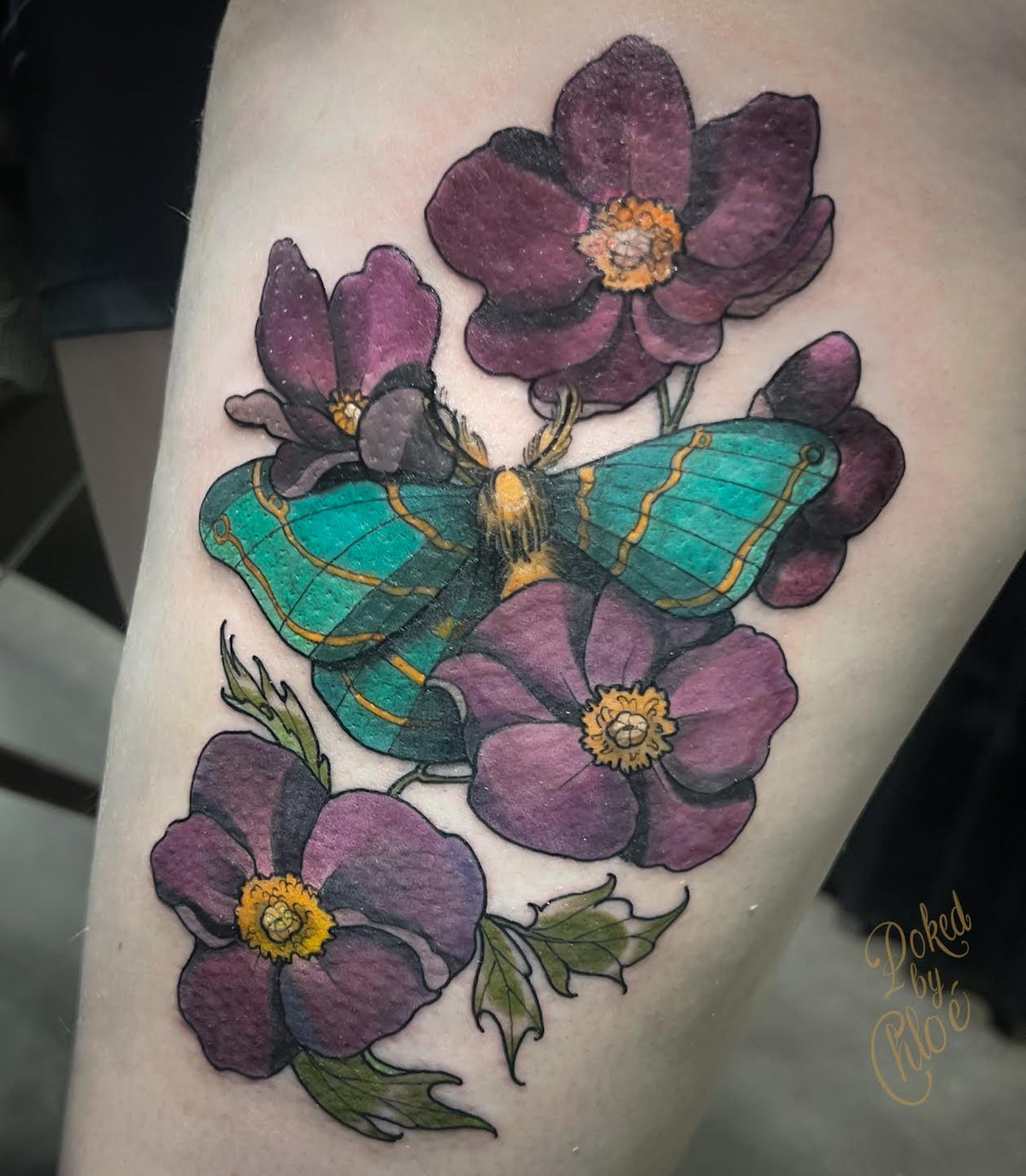 A close-up of a tattoo on skin showing a green and teal moth with yellow accents surrounded by dark purple flowers with yellow centers.