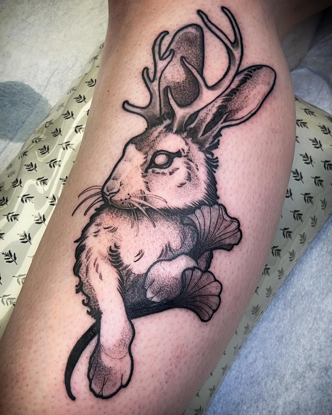 A black and gray hand-drawn style tattoo of a rabbit with antlers on a person's upper calf, shown against a tattoo sheet background.