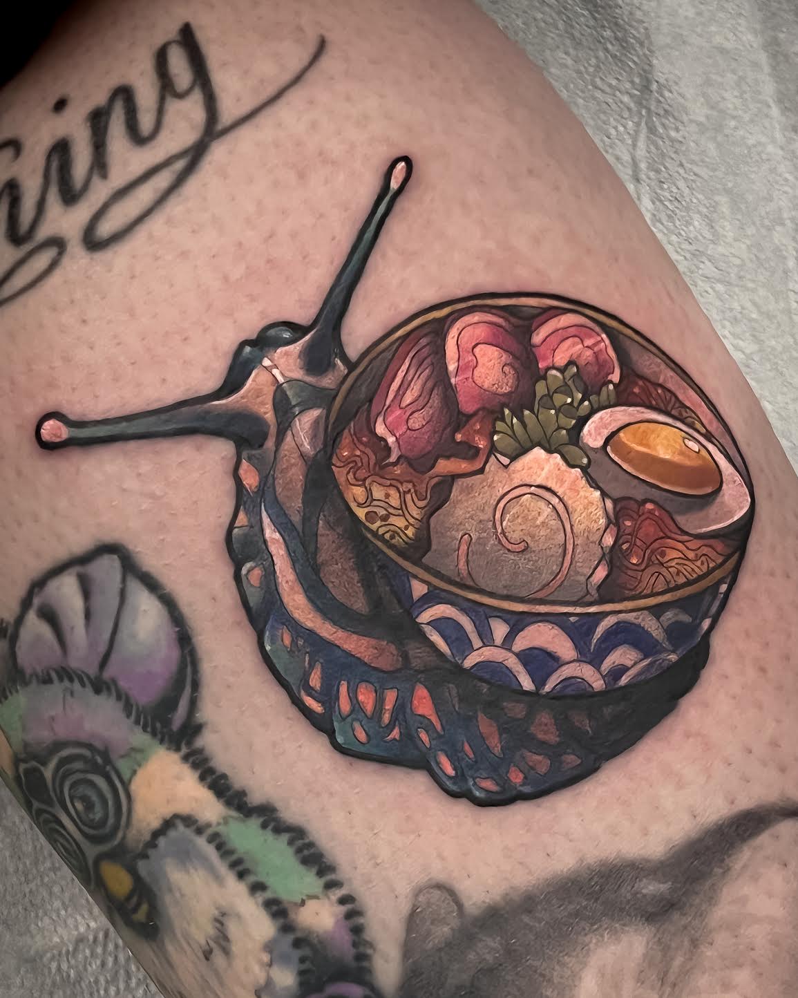 A close-up of a tattoo on human skin depicting a bowl of ramen with a snail-shaped handle and shell, surrounded by other tattoo artwork.
