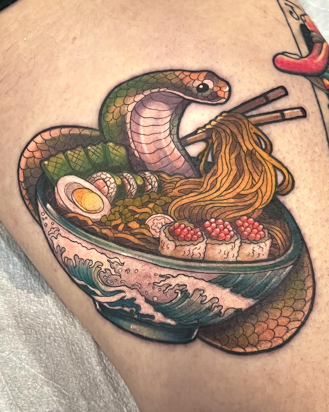 A close-up of a tattoo on human skin depicting a bowl of ramen with noodles, chopsticks, a snake coiled over the bowl, sushi, and wave-style detailing in black, green, orange, pink, and blue ink.