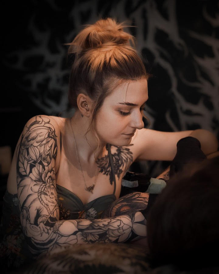 A woman with intricate black floral and tribal tattoos on her arms and chest, wearing a dark green strap top, captured in a dimly lit setting with a dark patterned background.